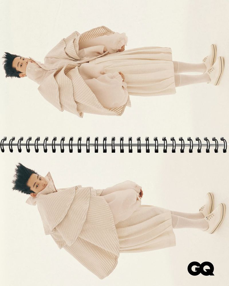 Editorial fashion shot of a person wearing a light tan layered outfit lying in a spiral notebook, featured in GQ magazine.