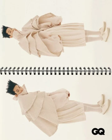 Editorial fashion shot of a person wearing a light tan layered outfit lying in a spiral notebook, featured in GQ magazine.