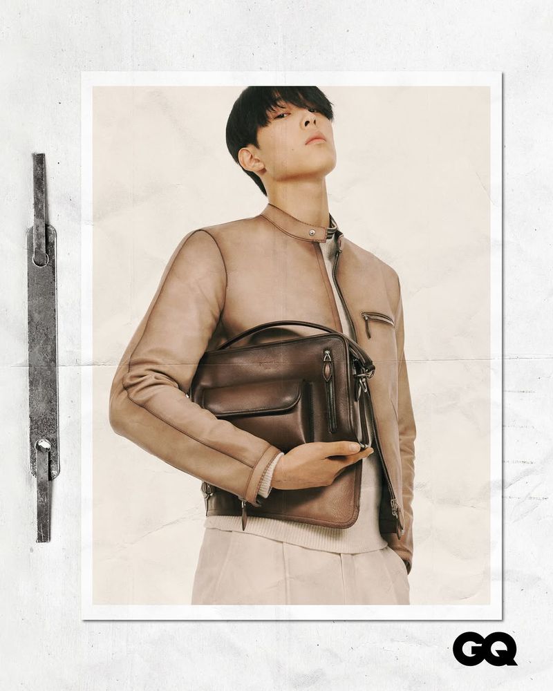 Handsome male fashion model poses in a neutral color leather jacket holding a dark brown leather briefcase in a studio fashion shot.