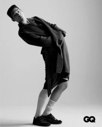Man wearing a dark coat, skirt, white socks, and dark shoes leaning backward against a grey seamless background