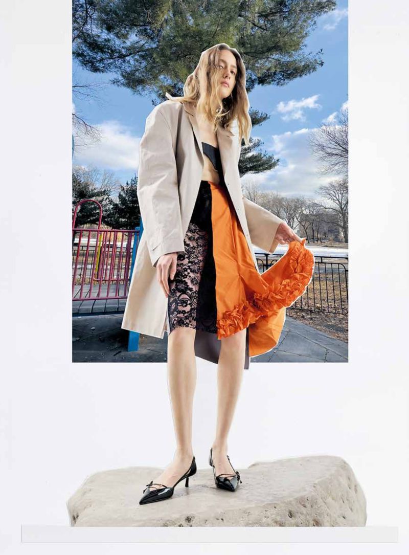 Fashionable young woman poses in front of a backdrop of a park on a stone platform, wearing a trench coat and unique layered skirt.