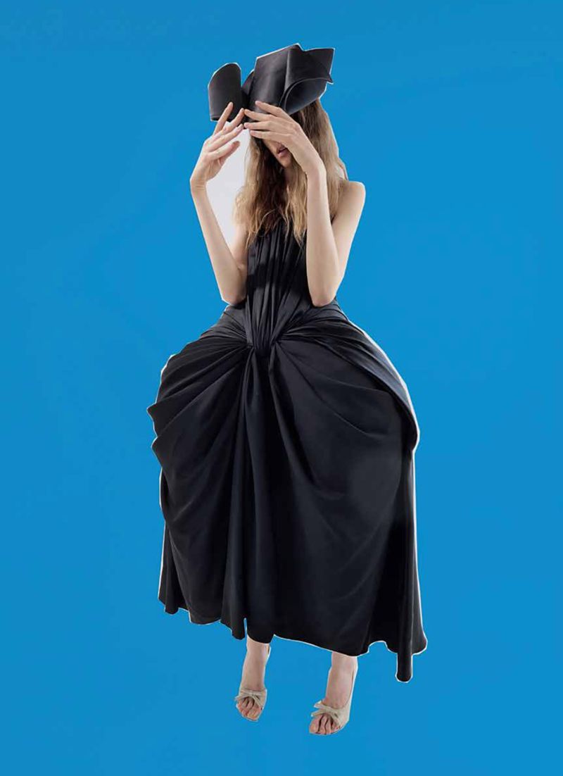 Fashion shot of a young adult woman in a black dress and dramatic headpiece, hands raised, against a bright blue backdrop. 