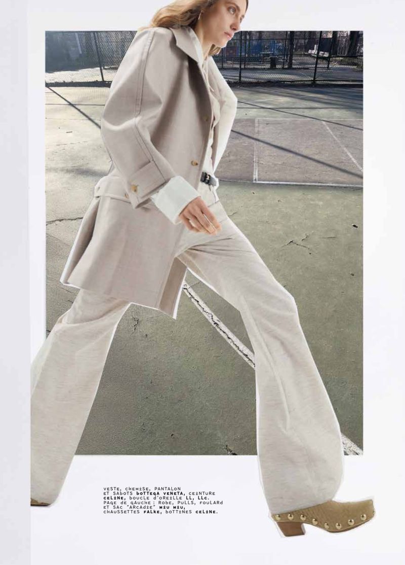 Fashion editorial shot on a tennis court: A long-haired model in a tan belted trench coat with white pants and clog shoes strides forward.