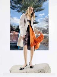 Model Lia Pavlova in a beige coat and orange-ruffled skirt standing on a rock against a park background for Mixte.