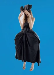 Model in a sculptural black dress and unique headwear posing against a solid blue background for Mixte.