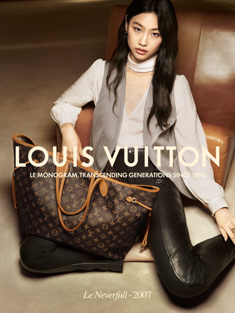 Brunette woman poses with Louis Vuitton handbag indoors, wearing a white blouse and gray vest, seated in tan leather chair.