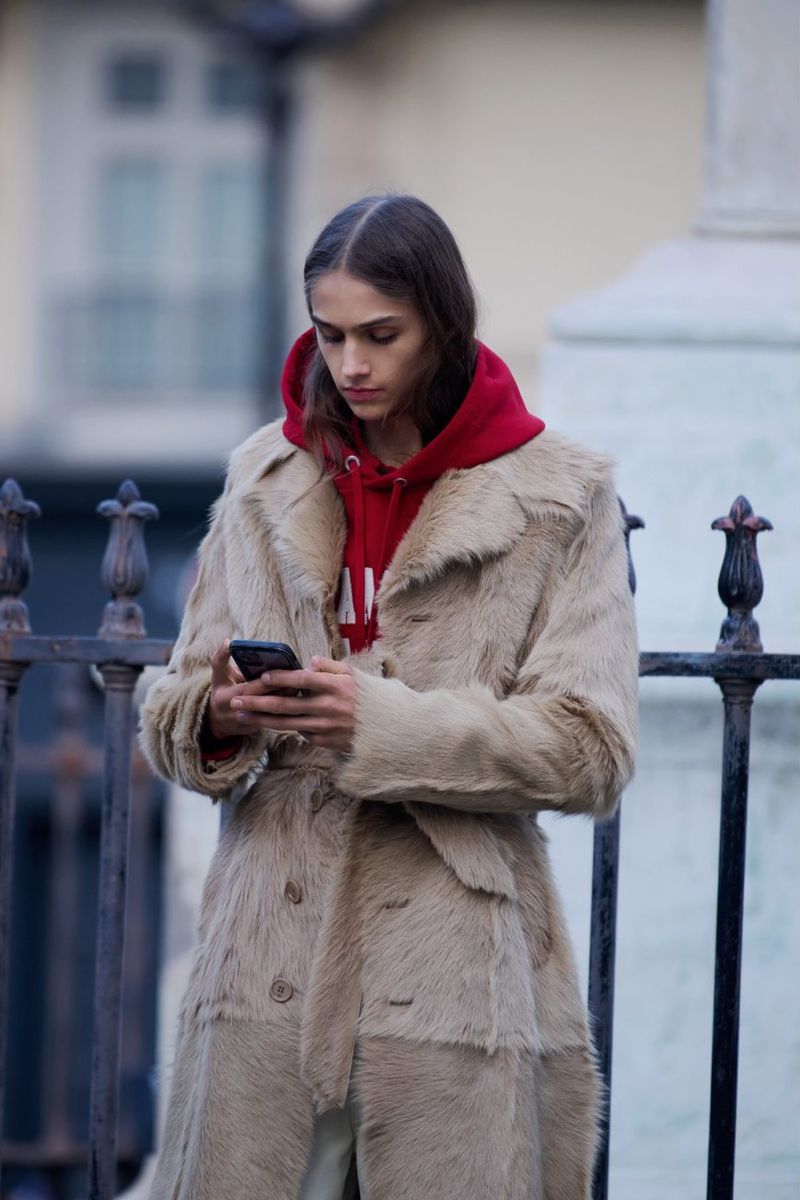 A woman in a fur coat and red hoodie uses a smartphone in an outdoor urban setting, framed by decorative wrought-iron fencing.
