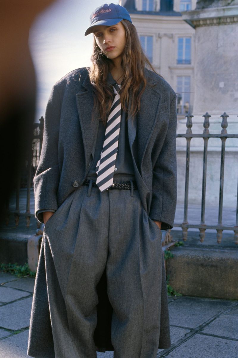 Model wearing a grey oversized coat, trousers, striped tie, and blue cap standing outdoors on a paved street.