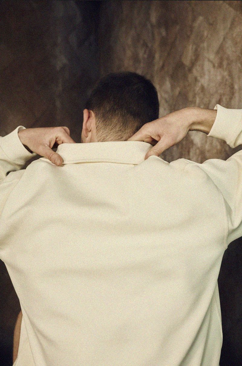 An adult wearing a cream-colored sweater stands against a textured brown backdrop adjusting the collar, facing away from the viewer.