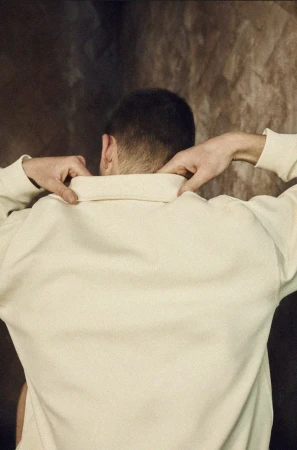 An adult wearing a cream-colored sweater stands against a textured brown backdrop adjusting the collar, facing away from the viewer.