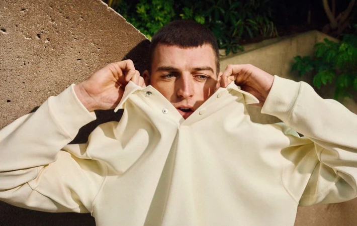 Close-up portrait of an adult man with short dark hair, wearing a pale yellow collared shirt, gazing into the distance with a neutral expression.