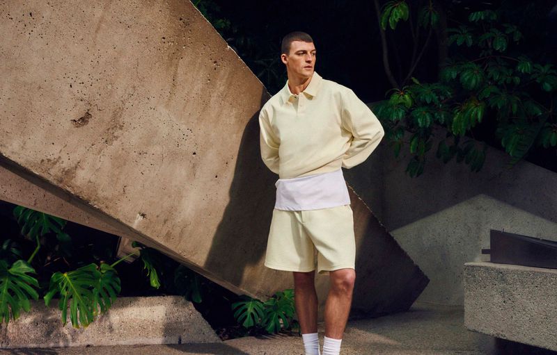Man in a pale yellow matching top and shorts stands in front of a concrete wall with dark green foliage in the background.