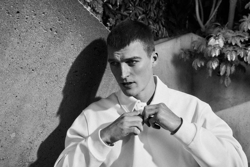 Portrait of a young adult male with short dark hair fastening the top button of a white collared shirt in an outdoor setting.