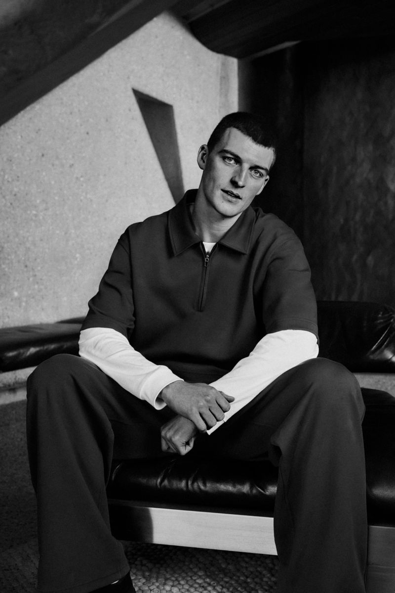 Monochrome portrait of a man in a red two-piece outfit, sitting on a black leather chair against a textured wall with architectural shadows.