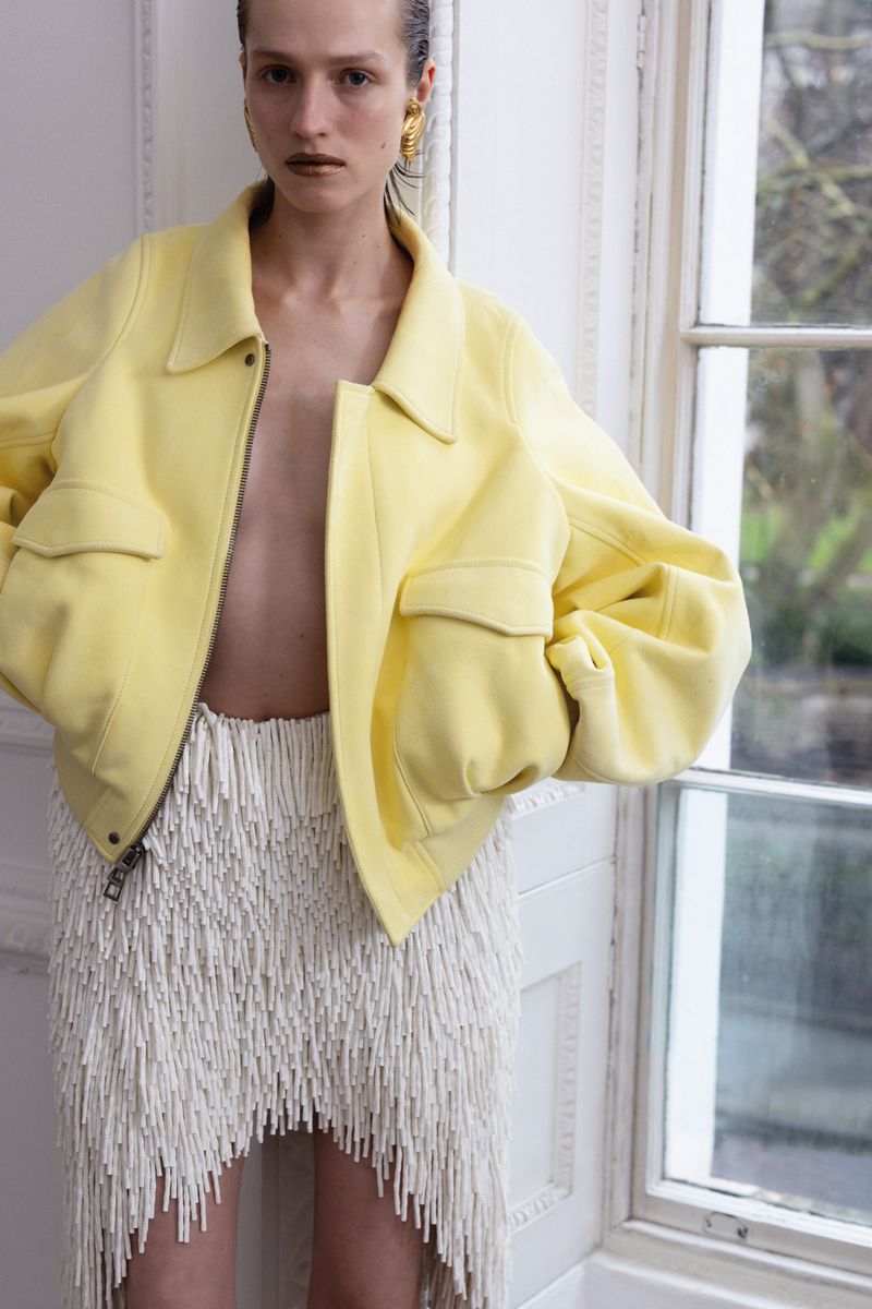 Indoor fashion shot of a posing model wearing a yellow jacket and white skirt with tassels, standing in front of a window.
