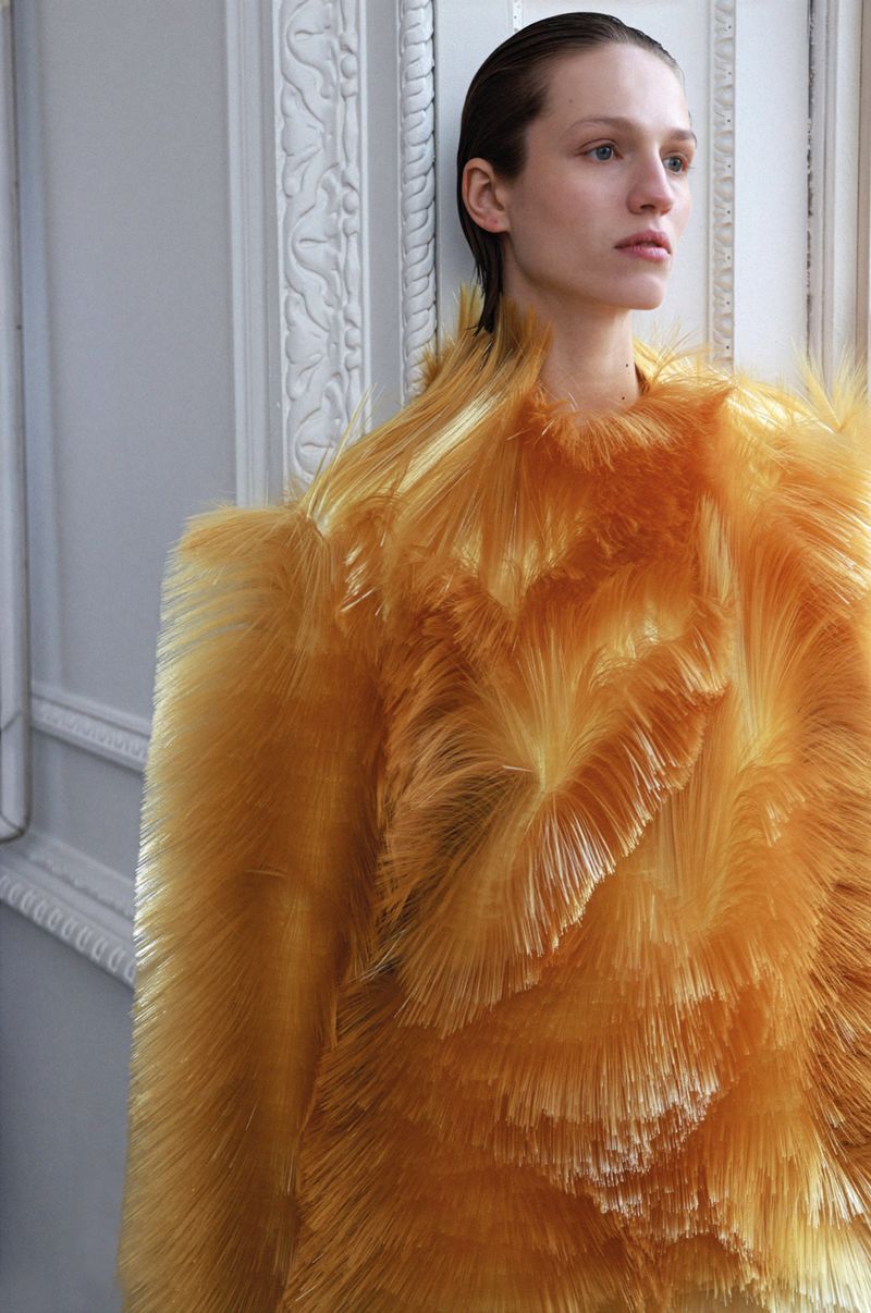 Fashion shot of an adult woman with wet hair wearing a textured, light-orange, sculptural top standing indoors against an ornate white wall.