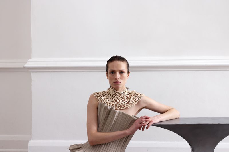 Fashion shoot of a woman in a beige dress with a detailed collar, resting her hands on a dark table against a white wall backdrop.