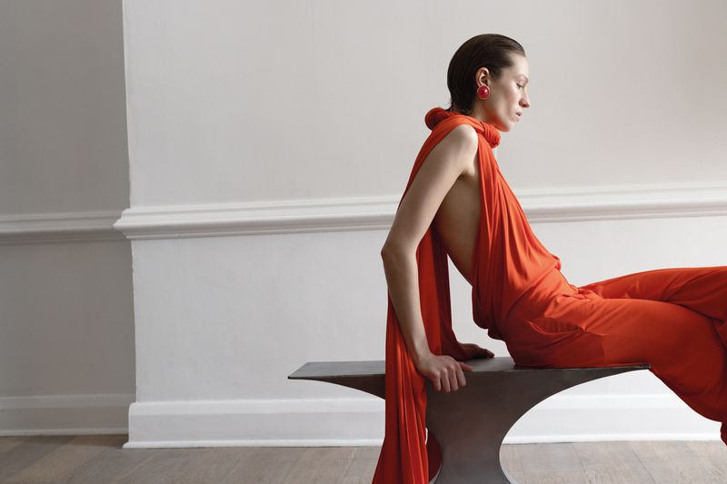 Woman in orange dress sits on metal bench in minimalist setting, showcasing fashion in a studio-lit environment, emphasizing color and style.