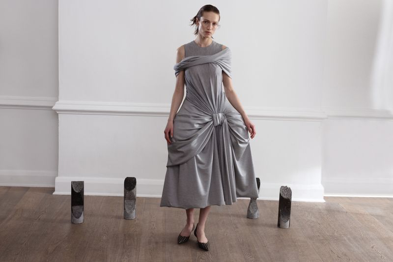 A fashionable woman in a silver dress poses in front of a white wall with decorative stone objects placed on a light brown floor.