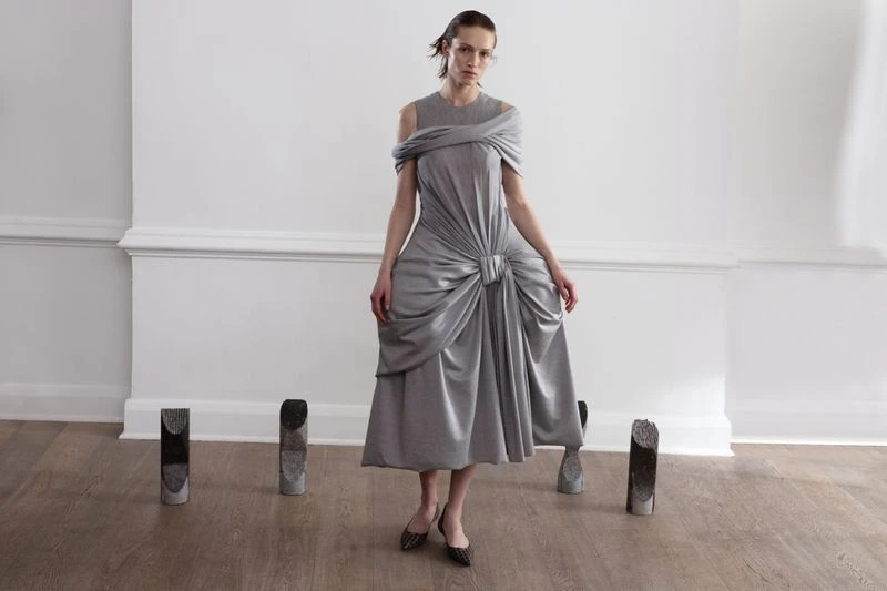A fashionable woman in a silver dress poses in front of a white wall with decorative stone objects placed on a light brown floor.