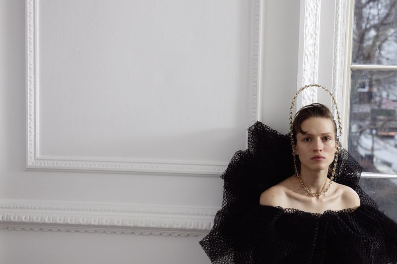 Woman with artistic headpiece and black ruffled collar posing in a white room near window light, fashion photography, looking at the camera.