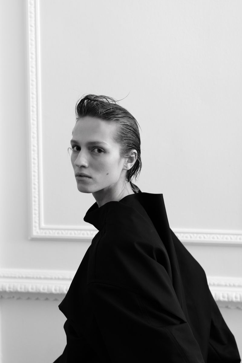 Monochrome portrait of a young adult wearing a black top against a white wall with ornate moulding, wet hair, and a neutral expression.