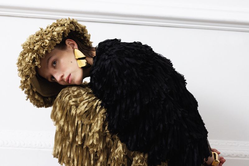 Fashion portrait of a person wearing a black and gold fringed jacket and hat with chunky gold earrings posing in a bright indoor space.