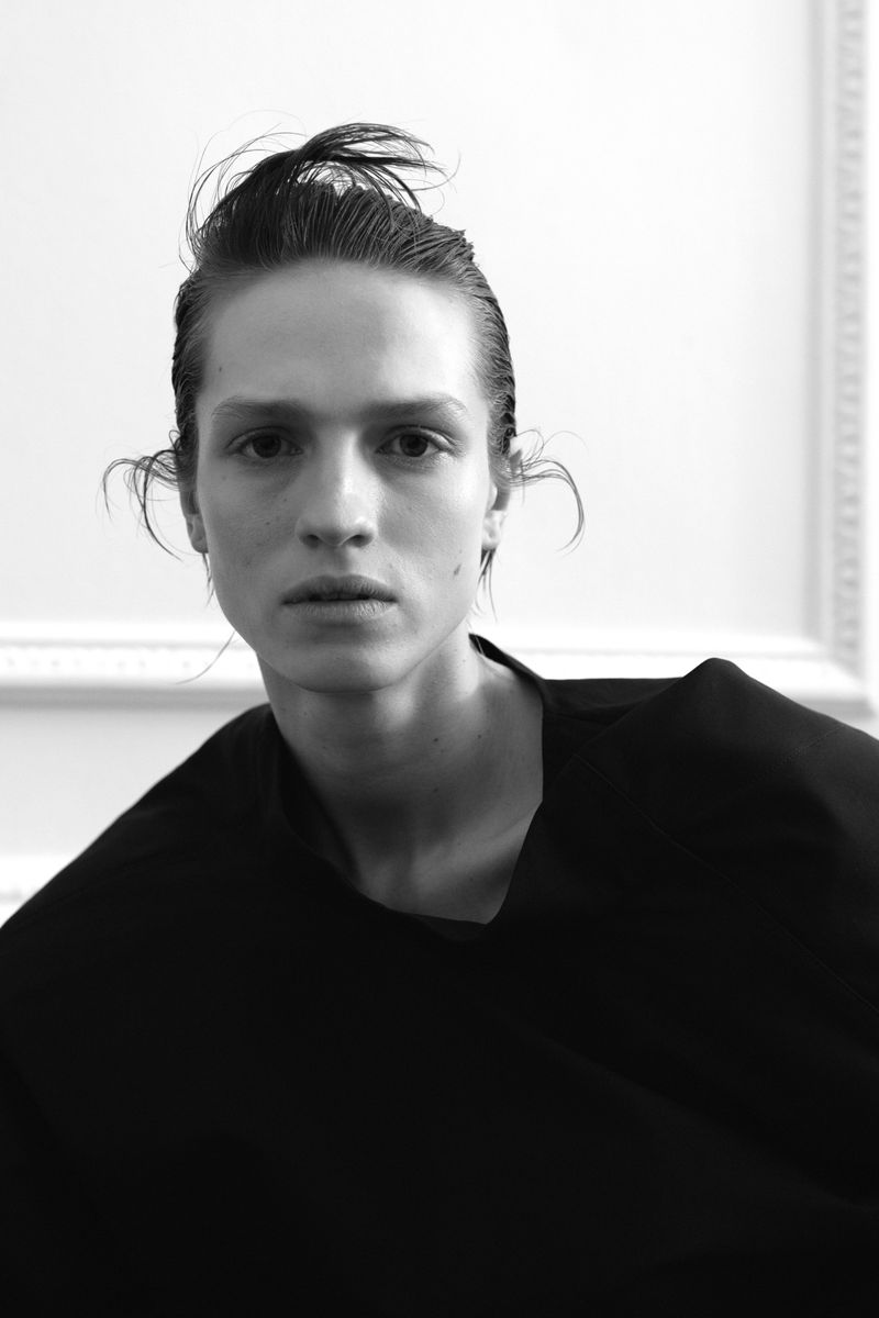 Black-and-white portrait of a young adult with slicked-back hair, wearing a black top against a plain white wall with moulding.