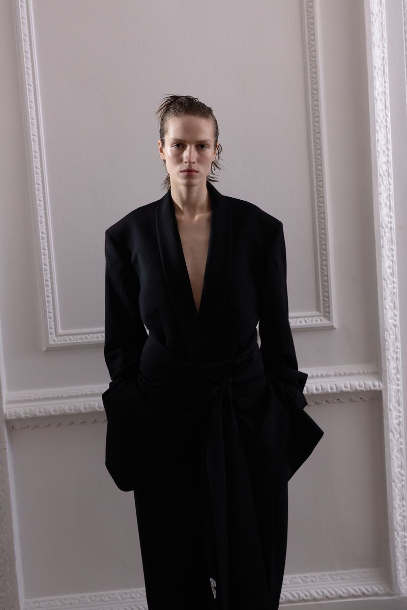 Woman in a draped black dress stands in front of a neutral wall with framed moulding in a fashion shot with wet-look hair.