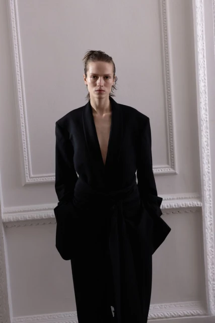 Woman in a draped black dress stands in front of a neutral wall with framed moulding in a fashion shot with wet-look hair.