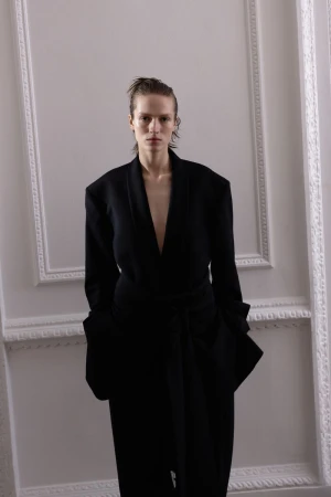 Model wearing a black tailored coat against white architectural wall molding in a portrait for The Blend Magazine
