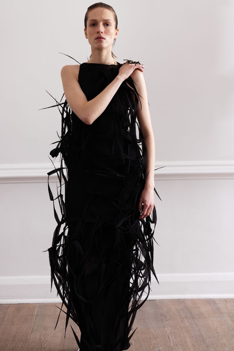 A slender adult female is shown indoors, facing forward, wearing a long black feather dress against a plain white wall in portrait framing.