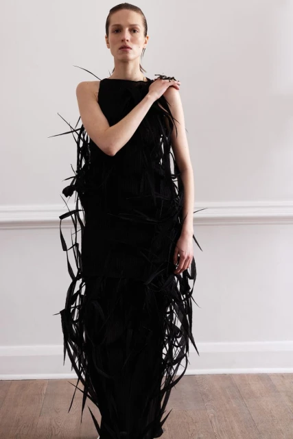 A slender adult female is shown indoors, facing forward, wearing a long black feather dress against a plain white wall in portrait framing.