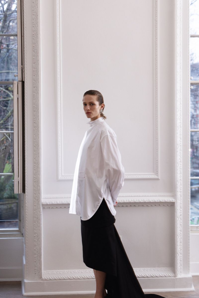 A woman in a white shirt and black skirt poses in a room with large windows and white walls, creating a minimalist fashion shot.