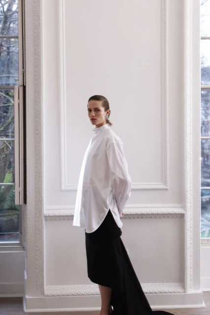A woman in a white shirt and black skirt poses in a room with large windows and white walls, creating a minimalist fashion shot.