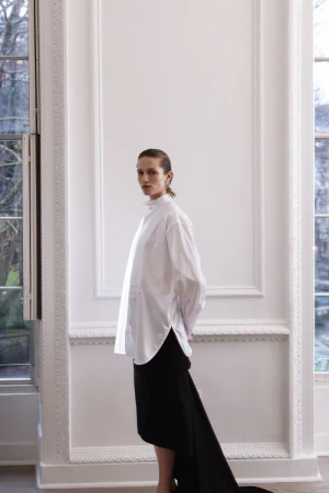 A woman in a white shirt and black skirt poses in a room with large windows and white walls, creating a minimalist fashion shot.