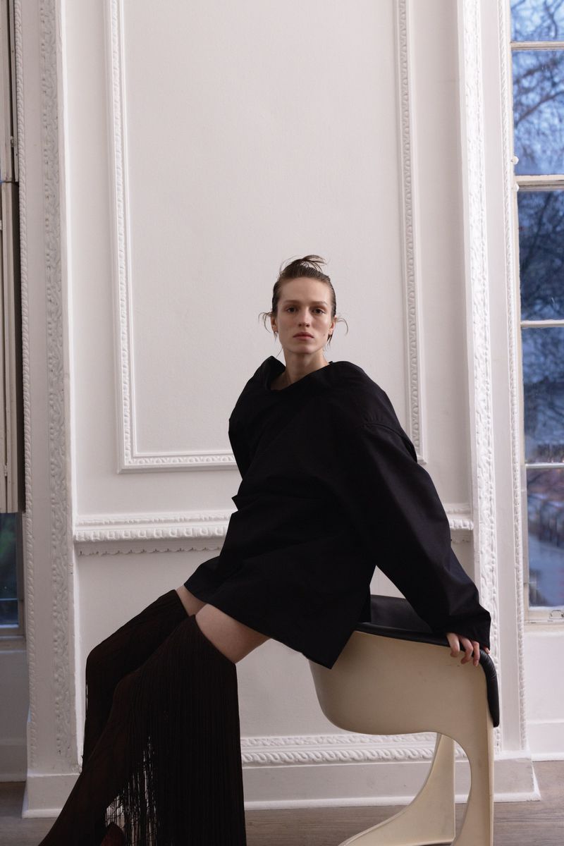 Fashion shot of a posed woman in a black top and fringe thigh-highs sitting on a chair in front of a white paneled wall with a window.