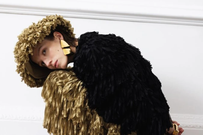 Model wearing a textured gold hat and jacket with black ruffles against a plain white wall