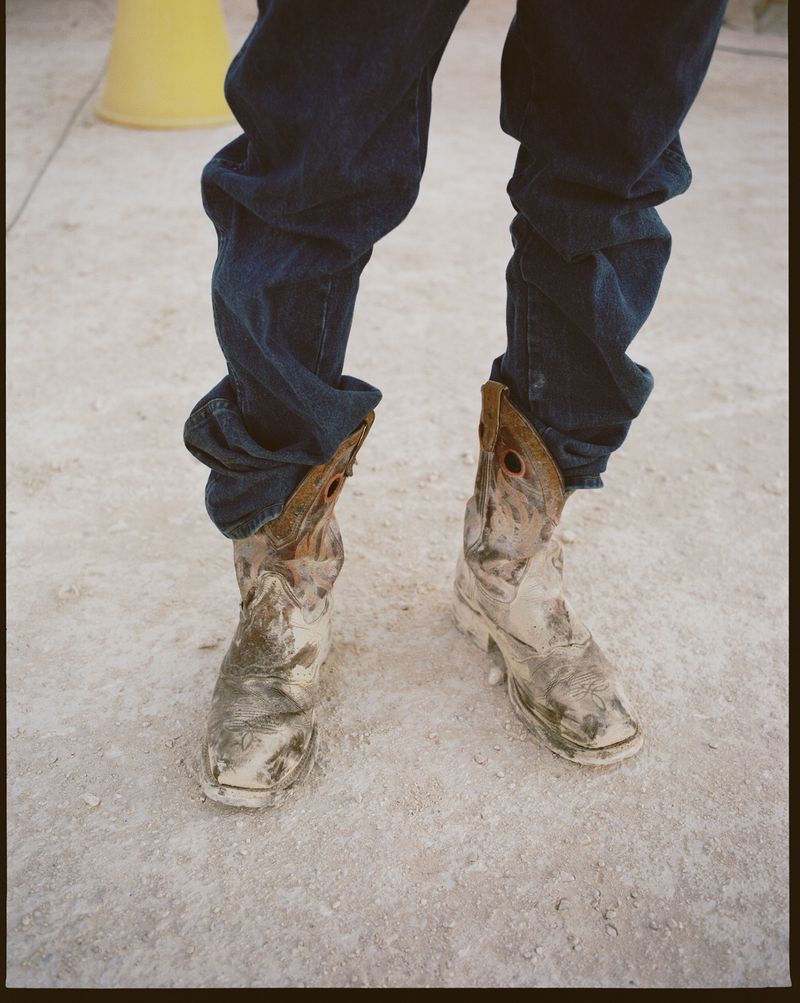 Close-up of a person wearing dark blue jeans and worn, light-colored cowboy boots on a textured, light-colored surface in an outdoor setting.
