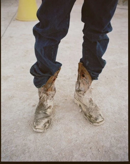 Close-up of a person wearing dark blue jeans and worn, light-colored cowboy boots on a textured, light-colored surface in an outdoor setting.