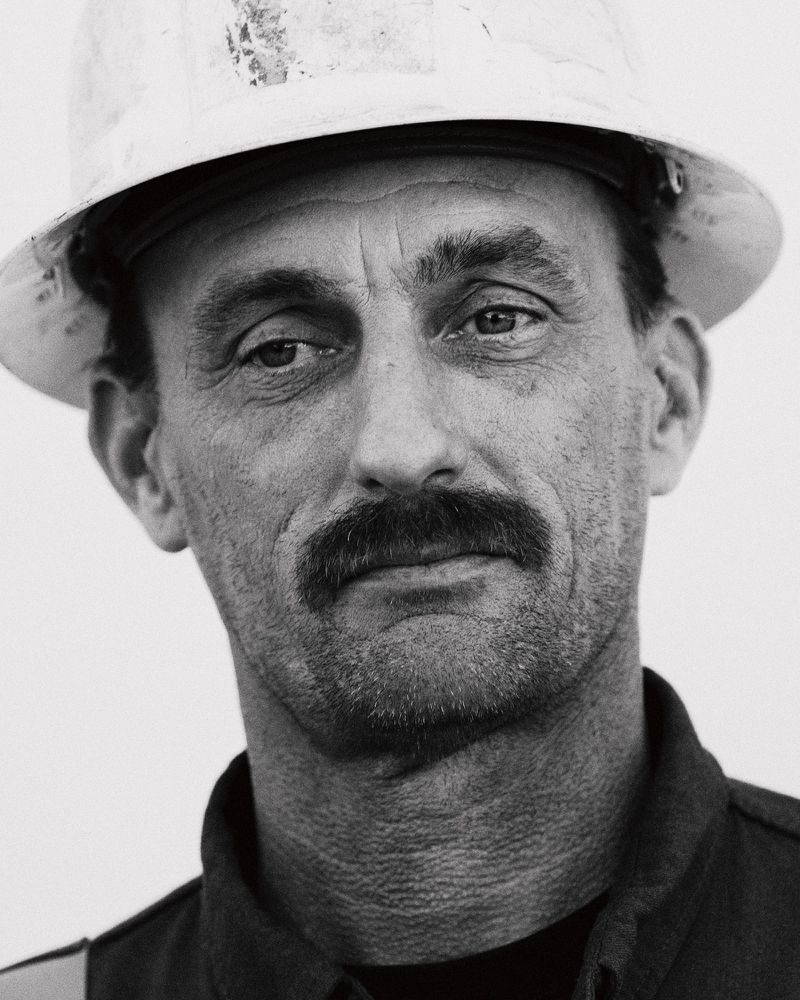 Monochrome portrait of a construction worker with a mustache wearing a hard hat against a plain white backdrop, medium close-up.