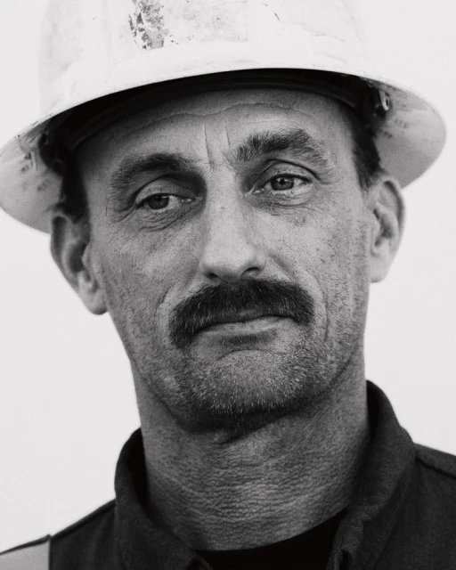 Monochrome portrait of a construction worker with a mustache wearing a hard hat against a plain white backdrop, medium close-up.