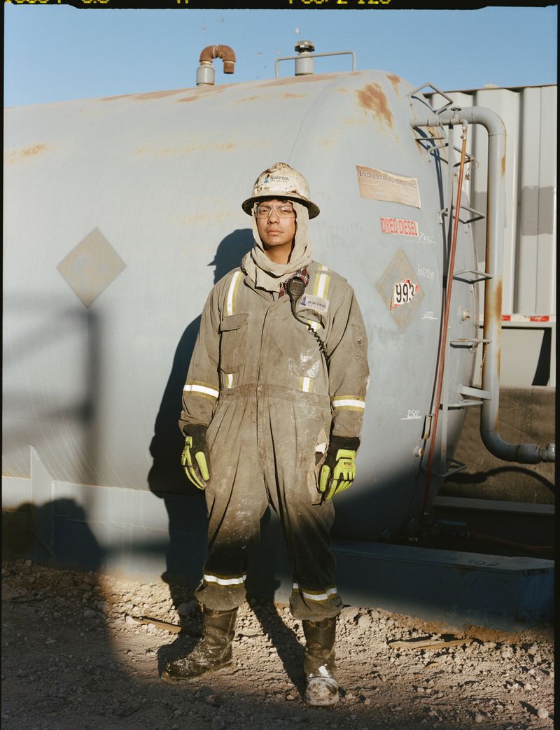 Man in work gear stands near a gray tank outdoors. He wears a hardhat, green gloves, and dark boots, with clear sky above.