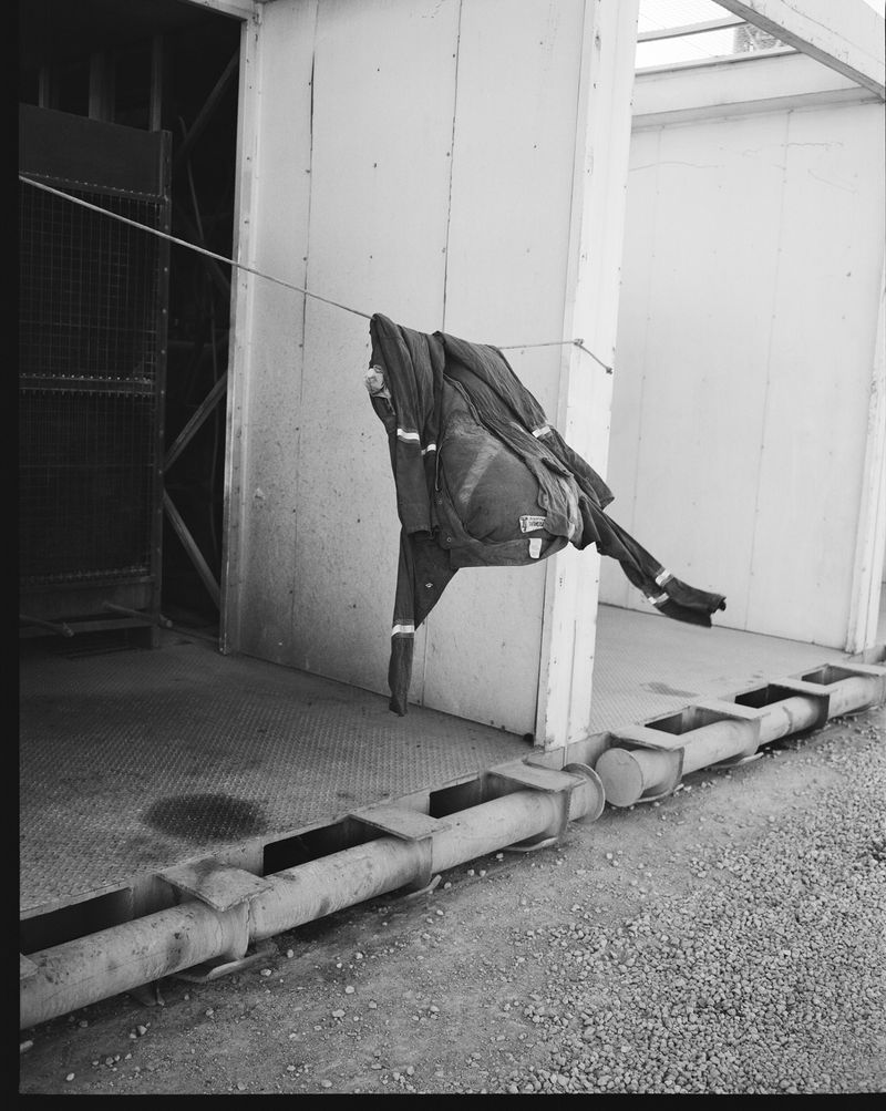 Black and white photo of a jacket hanging on a rope outdoors, with industrial pipes and gravel in the foreground.