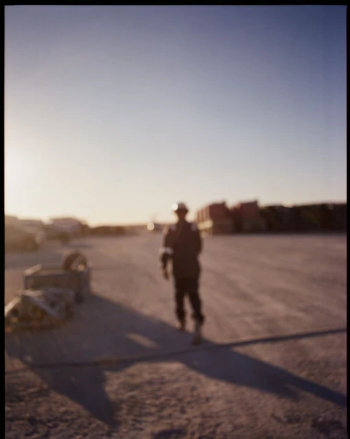 Worker in hard hat walking across a gravel lot at dusk, with industrial buildings and heavy equipment in the blurred background.