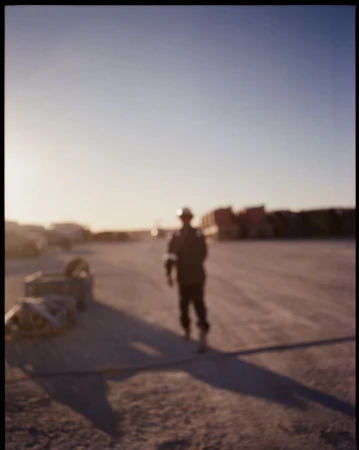 Worker in hard hat walking across a gravel lot at dusk, with industrial buildings and heavy equipment in the blurred background.