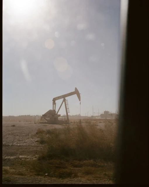 Pumpjack operating in a hazy, sunlit field under a blue sky. The pumpjack is viewed through an obstructed window, adding a framed perspective.
