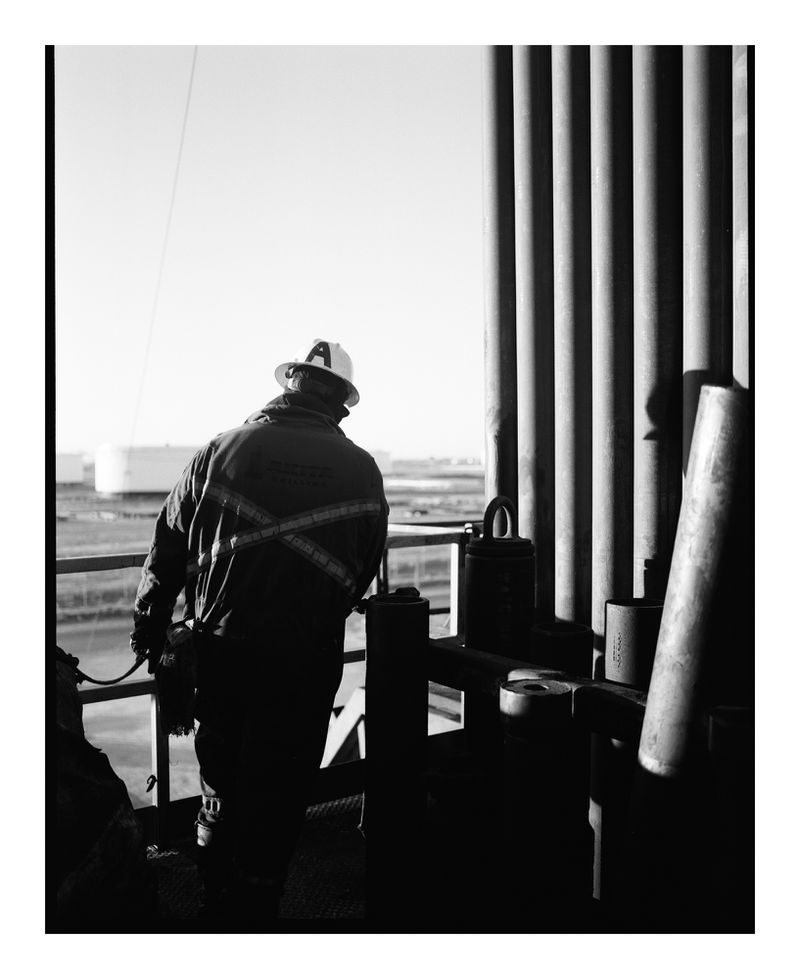 Monochrome shot of a worker in safety gear on an industrial platform, featuring metal pipes, tanks in the background, and a clear sky.