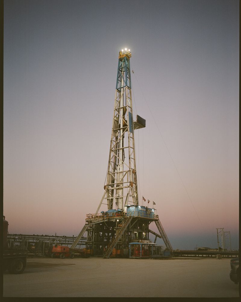 Tall oil drilling rig structure against the sky at dusk. Towering metal framework with lights on top. Industrial machinery in open setting.