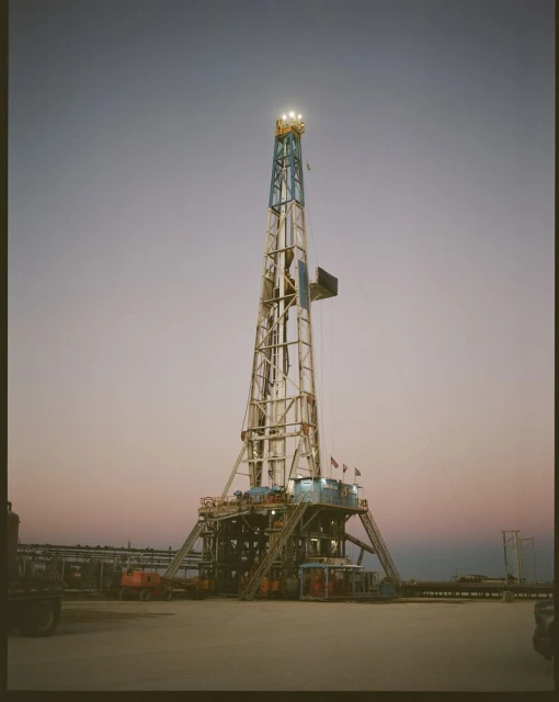 Tall oil drilling rig structure against the sky at dusk. Towering metal framework with lights on top. Industrial machinery in open setting.